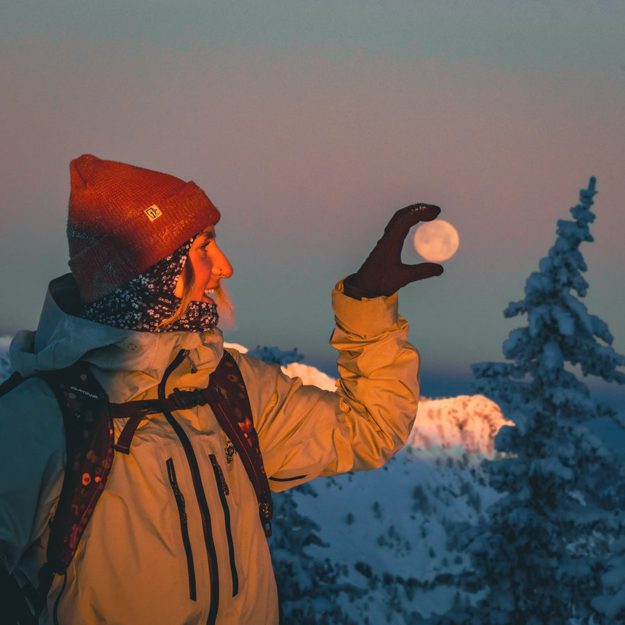 Clad in winter attire and sporting Icelantic Skis’ Maiden 102, a person appears to pinch the full moon between gloved fingers at sunset, framed by snow-covered trees and rugged mountain peaks.