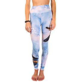 A woman stands barefoot, wearing Icelantic's 23/24 Soaring Kestrel Leggings—blue and pink cloud-patterned leggings featuring a feather and eagle design. Crafted from Hyper Wiking WIKMAX Fabric, these leggings offer a high-waist waistband for added comfort. Her hands rest relaxed by her sides, with a black bracelet adorning her left wrist.