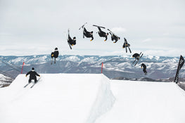 A skier rides the Icelantic Skis Nomad 94, performing a backflip over a snow ramp shown in multiple stages of motion, with snowy mountains in the background.