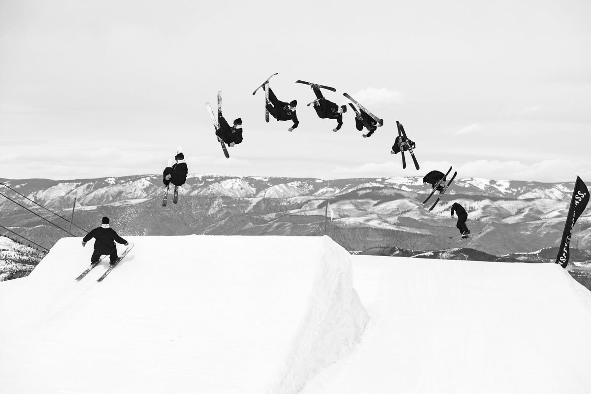 A skier performs a backflip off a snowy jump, captured in multiple stages of motion against a mountainous backdrop in black and white.