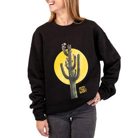 A smiling person with light brown hair stands with one hand in their pocket, wearing the Cactus Crew—a black unisex crewneck sweatshirt from Icelantic Skis featuring a large cactus and yellow sun graphic with the text "INTO THE WEST." This cozy style is made from recycled polyester fleece.