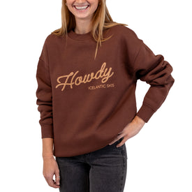 A smiling person wearing a brown Howdy Crew sweatshirt by Icelantic Skis—a cozy, unisex pullover crafted from recycled polyester sweater material—featuring “Howdy Icelantic Skis” in cursive on the front, paired with dark jeans and standing with one hand on their hip.