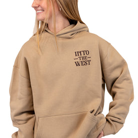 A smiling person wears a tan Cowboy Up Hoodie by Icelantic Skis, featuring "INTO THE WEST" printed on the front left side and made from recycled polyester, standing against a white background.