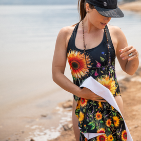 A woman in a sunflower swimsuit stands by the water, holding the Icelantic Skis Sunflower Travel Towel. She wears a black cap, a beaded necklace, and her hair is wet. The beach and lake are visible in the background.
