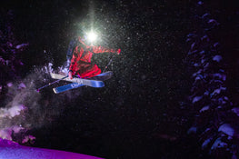 A skier outfitted in red gear, sporting a headlamp, soars mid-air on Icelantic Skis’ Maiden 94 as snow sprays around them, illuminated against the dark, snow-laden forest backdrop.