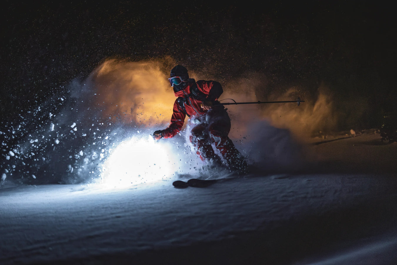 A skier on Icelantic Skis’ Maiden 102 carves through snow at night, illuminated by a bright light, with red gear, a spray of snow, and a dark background creating a dramatic effect.