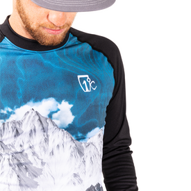 A man wearing a gray hat and the Icelantic 23/24 Mens WikMax Baselayer Top, which features a mountain and sky design from the Pioneer Collection, looks down. The image captures part of his face, chest, and shoulder against a white background.