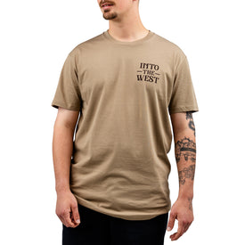 Man wearing a beige Icelantic Cowboy Up Tee, made from 100% combed cotton with INTO THE WEST text on the chest, black pants, and visible arm tattoos, standing against a white background.