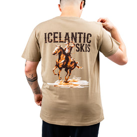 A person faces away, wearing the Icelantic Cowboy Up Tee—a tan t-shirt made from 100% combed cotton, featuring “Icelantic Skis” and an illustration of a rider on a rearing horse printed on the back. The person holds up the shirt’s collar with one hand, highlighting this unique design.