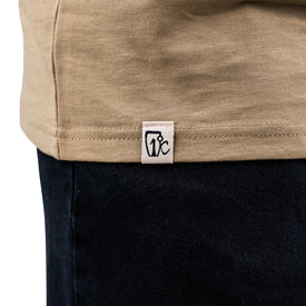 Close-up of a person wearing the Cowboy Up Tee by Icelantic—an artwork t-shirt crafted from 100% combed cotton—in beige, with a small white fabric tag at the hem featuring black logo or text, paired with dark blue denim jeans.