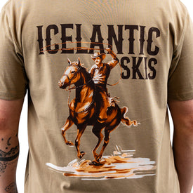 A person wearing a tan Cowboy Up Tee by Icelantic, a 100% combed cotton t-shirt featuring "ICELANTIC SKIS" and an image of a cowboy on horseback holding a lasso on the back.
