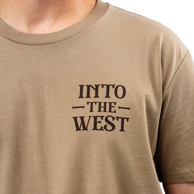 A person wearing a tan Cowboy Up Tee by Icelantic, crafted from 100% combed cotton, featuring the text INTO THE WEST printed in bold, dark letters on the left chest. The upper part of the person's torso is visible.