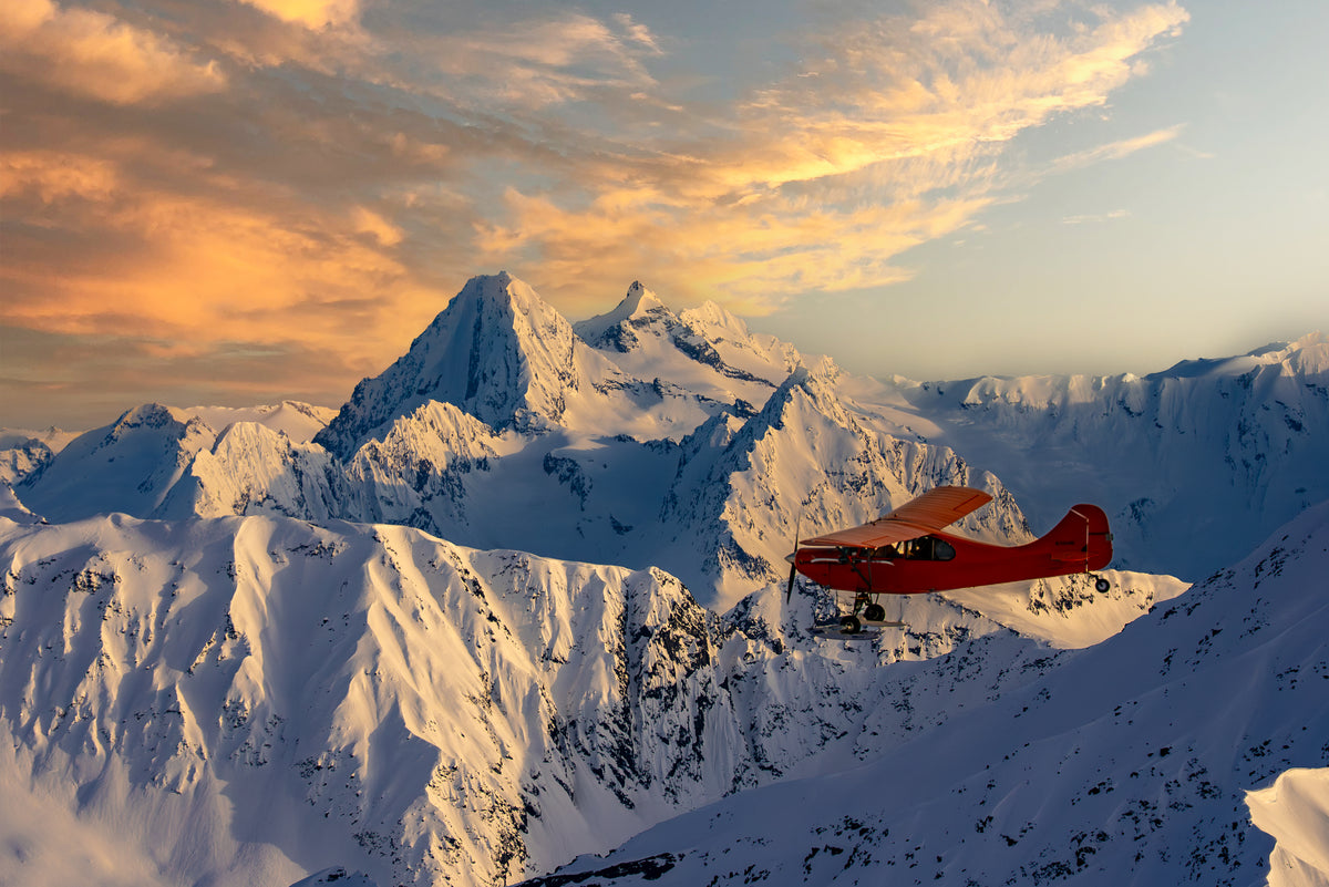 A red bush plane flies above snow-covered mountains at sunset, with dramatic clouds and golden light illuminating the peaks.