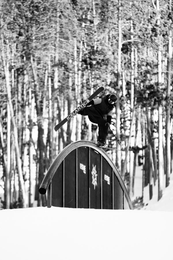 Balanced atop a curved rail in a snow park, a skier glides on Icelantic Skis’ Nomad 94, ski poles in hand and blurred trees forming the monochrome background.