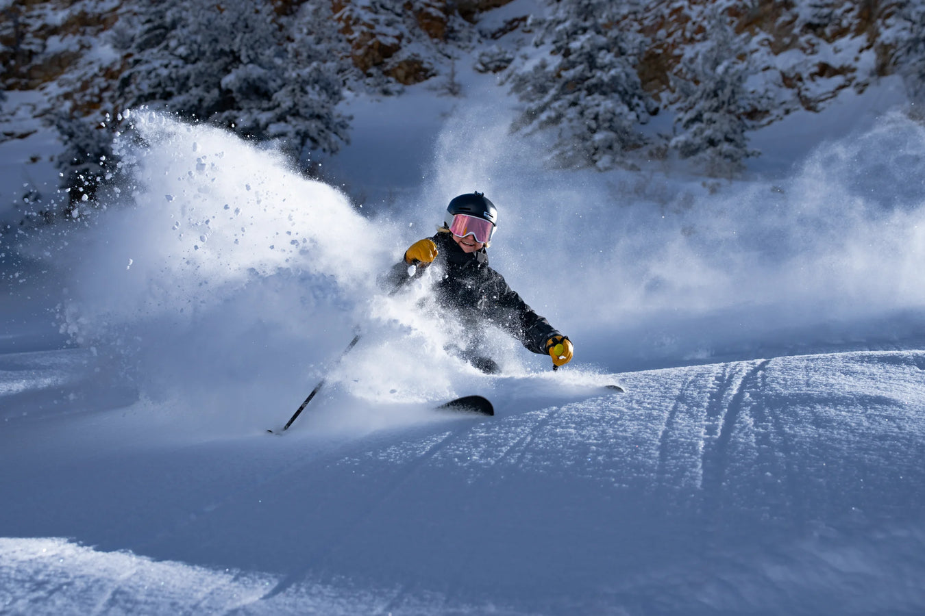 Wearing a helmet and goggles, a skier on Icelantic Skis’ Maiden 108 carves through fresh powder snow on a snowy slope, sending up a spray of snow, with snow-covered trees in the background.