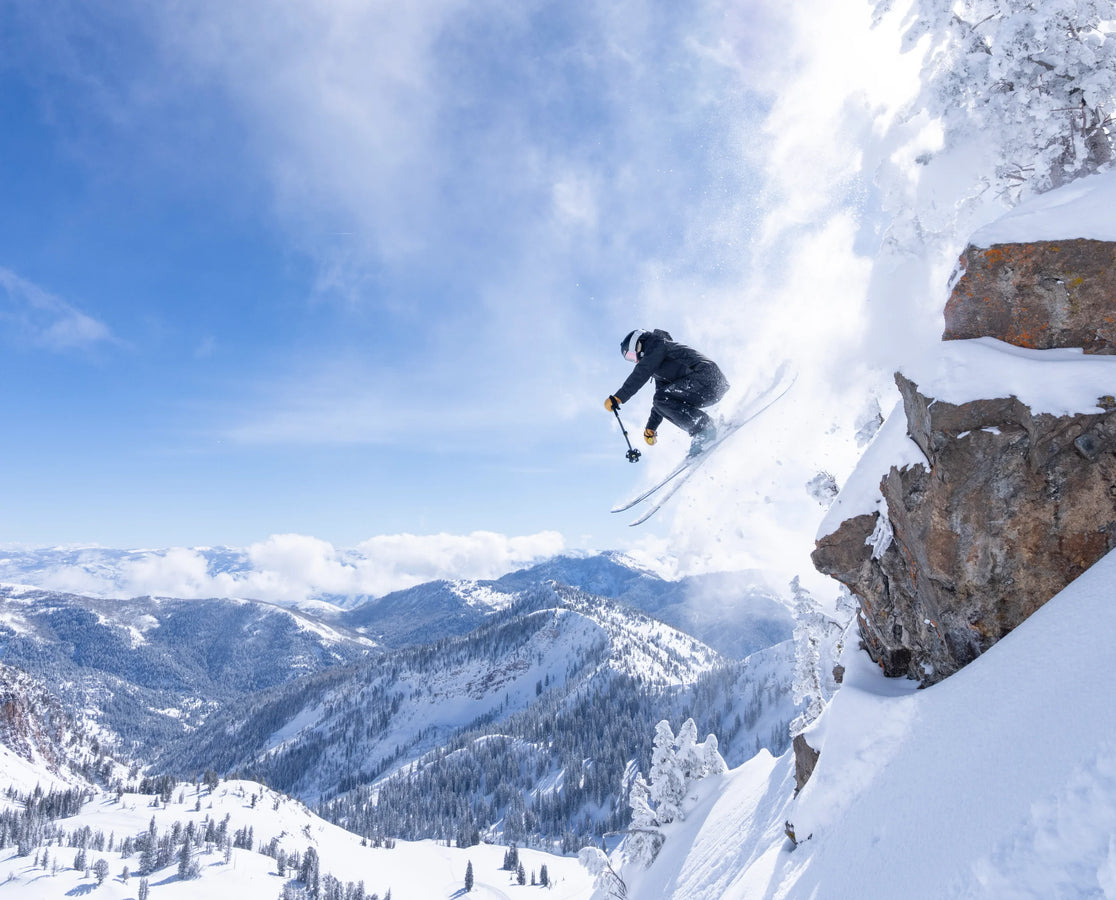 Soaring off a snowy cliff on the Maiden 108 by Icelantic Skis, a skier catches air amidst powder and brilliant sunlight, framed by snow-covered mountains and trees under a clear blue sky.