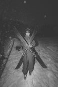 A skier dressed in winter gear stands in the snow at night, holding two crossed Icelantic Skis Nomad 94 models in front of them as snow falls and trees appear in the darkened background.