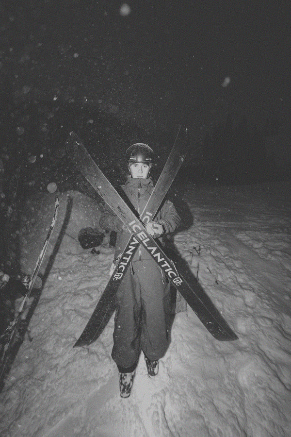 A skier dressed in winter gear stands in the snow at night, holding two crossed Icelantic Skis Nomad 94 models in front of them as snow falls and trees appear in the darkened background.