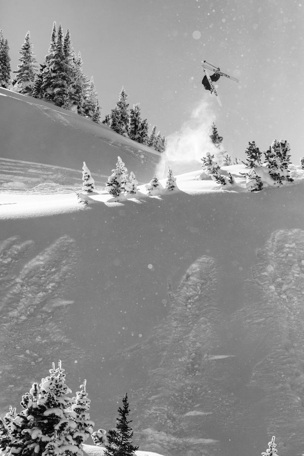A skier on Icelantic Skis’ Nomad 106 catches big air off a snowy slope, soaring above snow-covered pine trees beneath a clear sky.
