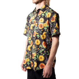 A person wearing the Icelantic Skis Sunflower Party Shirt—a short-sleeved, button-up shirt made from eco-friendly recycled polyester and featuring a colorful sunflower and foliage pattern inspired by Travis Parr artwork—stands against a white background.
