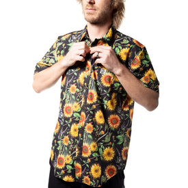 A person is buttoning up the Icelantic Skis Sunflower Party Shirt, a short-sleeve shirt made from eco-friendly recycled polyester and featuring a vibrant sunflower pattern inspired by Travis Parr’s artwork. The background is white.