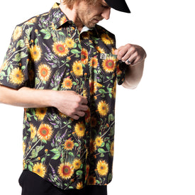 Wearing the Icelantic Skis Sunflower Party Shirt—an eco-friendly, short-sleeve shirt made from recycled polyester and adorned with a vibrant sunflower pattern—a person paired with a black cap looks down, adjusting or examining a small patch on the chest pocket against a white background.