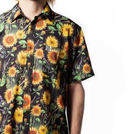 A person, shown from the waist up against a white background, is wearing the Icelantic Skis Sunflower Party Shirt—a black short-sleeve shirt made from eco-friendly recycled polyester and featuring a vibrant sunflower and floral pattern.