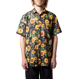 Wearing the Icelantic Skis Sunflower Party Shirt—a short-sleeve button-up featuring a vibrant sunflower and floral pattern made from eco-friendly recycled polyester—a person stands against a white background.