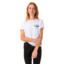 A woman stands against a white background, wearing the Icelantic Permit Tee—a GOTS certified organic cotton t-shirt featuring a small fish graphic and text on the left side—paired with black pants. She looks slightly to the side with a relaxed expression.