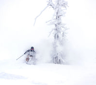 Wearing goggles and winter gear, a skier on Icelantic Skis’ Nomad 112 Whiteout carves through deep powder snow near a snow-covered tree, sending up a large cloud of snow.