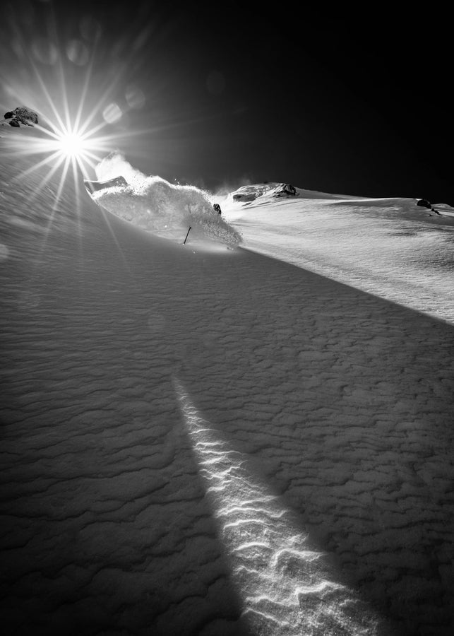 A black and white photo captures a skier carving down a snowy slope on Icelantic Skis’ Nomad 112 Whiteout, with the sun shining brightly overhead, casting striking shadows and illuminating powdery snow kicked up into the air.