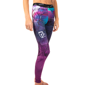 A woman stands barefoot against a white background, wearing the Icelantic 23/24 Womens WikMax Baselayer Full Bottoms featuring a colorful, abstract pattern in shades of purple, pink, and blue. The soft WIKMAX fabric displays a white Icelantic logo on the left thigh.