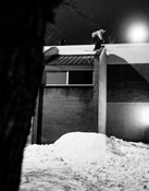A skier equipped with Icelantic Skis’ Nomad 94 Whiteout prepares to leap off a building ledge onto a snowy pile below at night in this black and white scene.