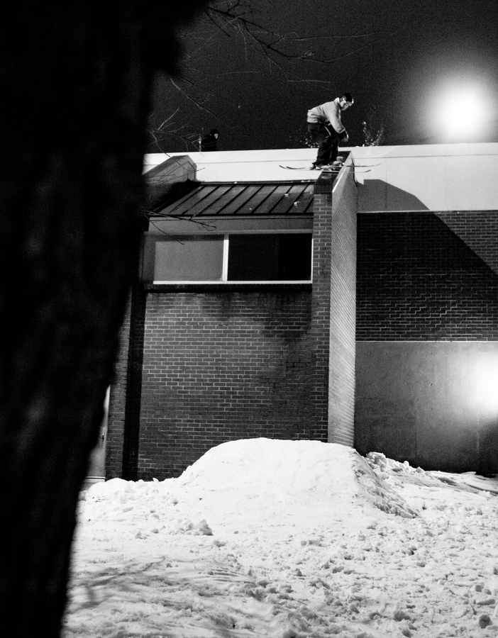 A skier equipped with Icelantic Skis’ Nomad 94 Whiteout prepares to leap off a building ledge onto a snowy pile below at night in this black and white scene.