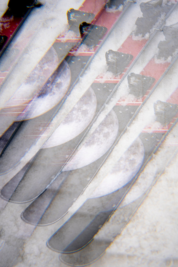 A close-up, double-exposed image of several Icelantic Skis Nomad 100 skis crossed over each other, featuring a moon graphic on their surfaces and a snowy background.