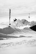 A skier on Icelantic Skis’ Nomad 94 performs a mid-air trick above a snowy slope, with mountains and ski lift cables visible in the cloudy background.