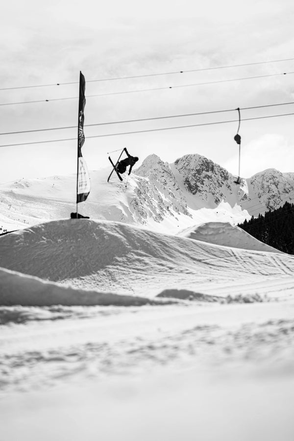 A skier on Icelantic Skis’ Nomad 94 performs a mid-air trick above a snowy slope, with mountains and ski lift cables visible in the cloudy background.