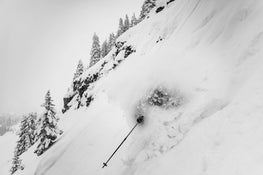 A skier equipped with the Icelantic Skis Nomad 112 Whiteout is nearly buried in deep powder on a snowy mountain slope, with only one ski pole visible amid snow-covered trees and rocks beneath a cloudy sky.
