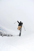 A skier launches into the air holding ski poles, riding Icelantic Skis’ Nomad 100 Whiteout, dressed in tan pants and a black jacket, with snow-covered trees and mountains in the background beneath a cloudy sky.