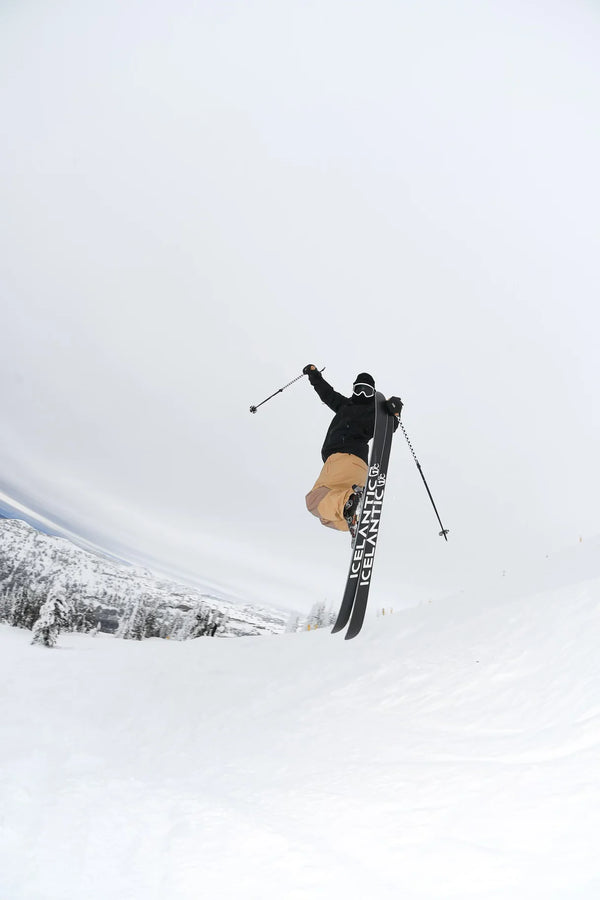 A skier launches into the air holding ski poles, riding Icelantic Skis’ Nomad 100 Whiteout, dressed in tan pants and a black jacket, with snow-covered trees and mountains in the background beneath a cloudy sky.