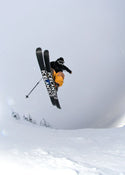 A skier wearing orange pants and a black jacket is mid-air performing a jump over snowy terrain with trees below, riding Icelantic Skis’ Nomad 100 Whiteout skis.