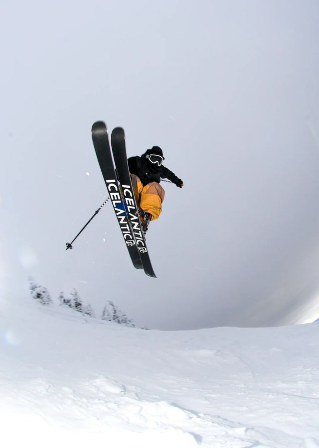 A skier wearing orange pants and a black jacket is mid-air performing a jump over snowy terrain with trees below, riding Icelantic Skis’ Nomad 100 Whiteout skis.