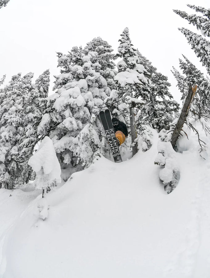 A skier in a yellow jacket launches off a snowy cliff among snow-covered trees, holding their Icelantic Skis Nomad 106 Whiteout mid-air against a cloudy winter sky.
