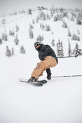 Wearing brown pants and a black jacket, a skier balances on one Icelantic Skis Nomad 106 while holding ski poles on a snowy mountain slope dotted with scattered trees in the background.