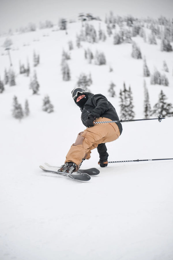 Wearing brown pants and a black jacket, a skier balances on one Icelantic Skis Nomad 106 while holding ski poles on a snowy mountain slope dotted with scattered trees in the background.