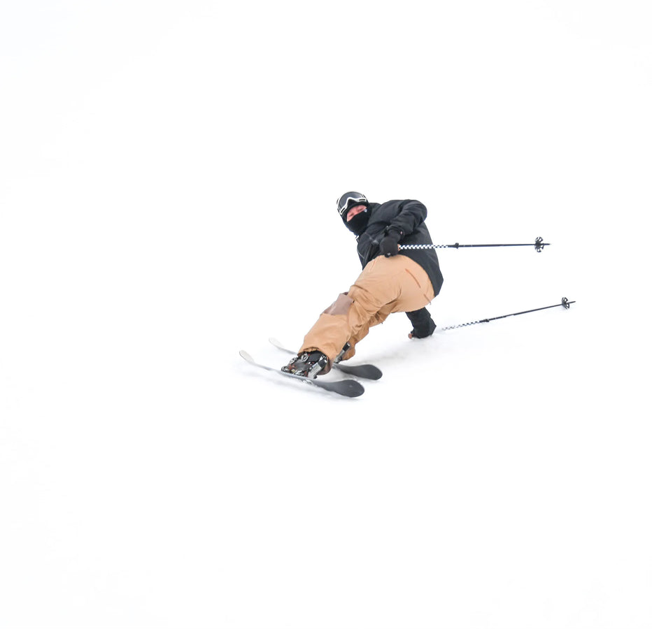 A skier carves a sharp turn on a snowy slope, leaning to one side with poles trailing behind, riding on Icelantic Skis’ Nomad 100 Whiteout against a pristine white backdrop.