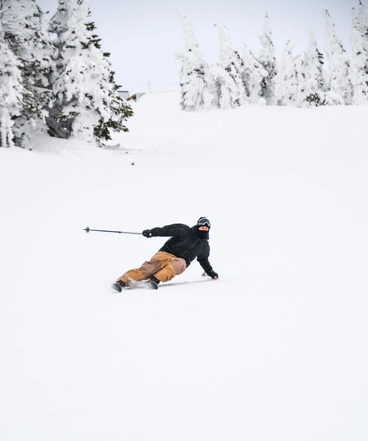 Wearing tan pants and a dark jacket, a skier carves down a snowy slope on Icelantic Skis’ Nomad 106, sending up a spray of powder among snow-covered trees with one pole outstretched.