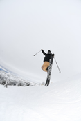 Wearing tan pants and a black jacket, a skier launches into a jump on a snowy slope with the Icelantic Skis Nomad 100, ski poles in hand and snow-covered trees and mountains forming the backdrop.