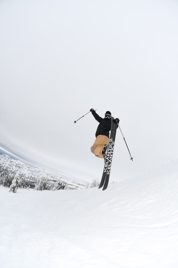 Wearing tan pants and a black jacket, a skier launches into a jump on a snowy slope with the Icelantic Skis Nomad 100, ski poles in hand and snow-covered trees and mountains forming the backdrop.