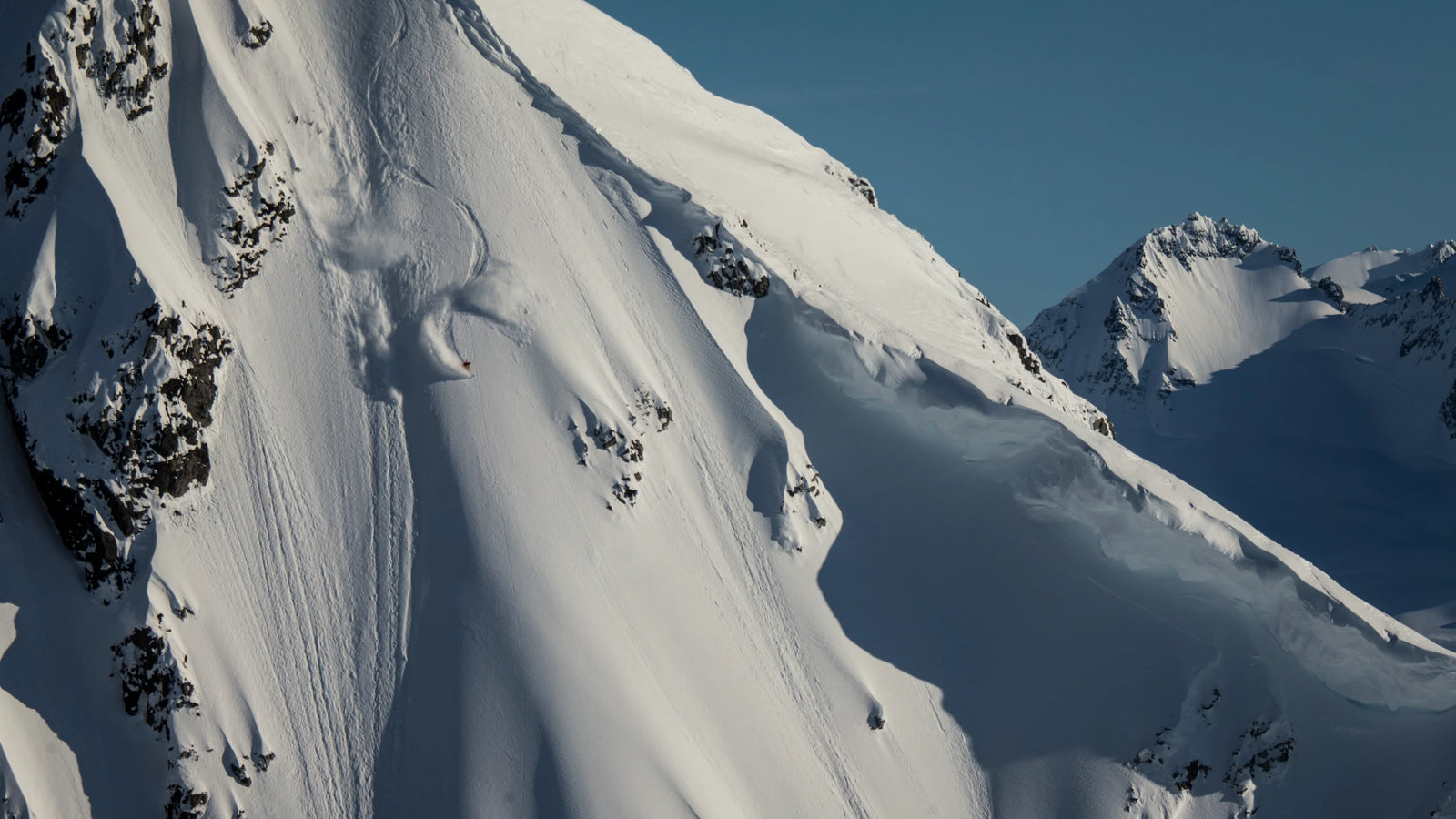 A skier descends a steep, snow-covered mountain slope on the Icelantic Skis Nomad 112, carving tracks in the fresh powder under clear blue skies with rugged snowy peaks rising in the background.